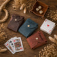 Three personalized leather card sleeves in brown, blue, and reddish-brown, styled on a rustic wooden table with playing cards, dice, and dried flowers.