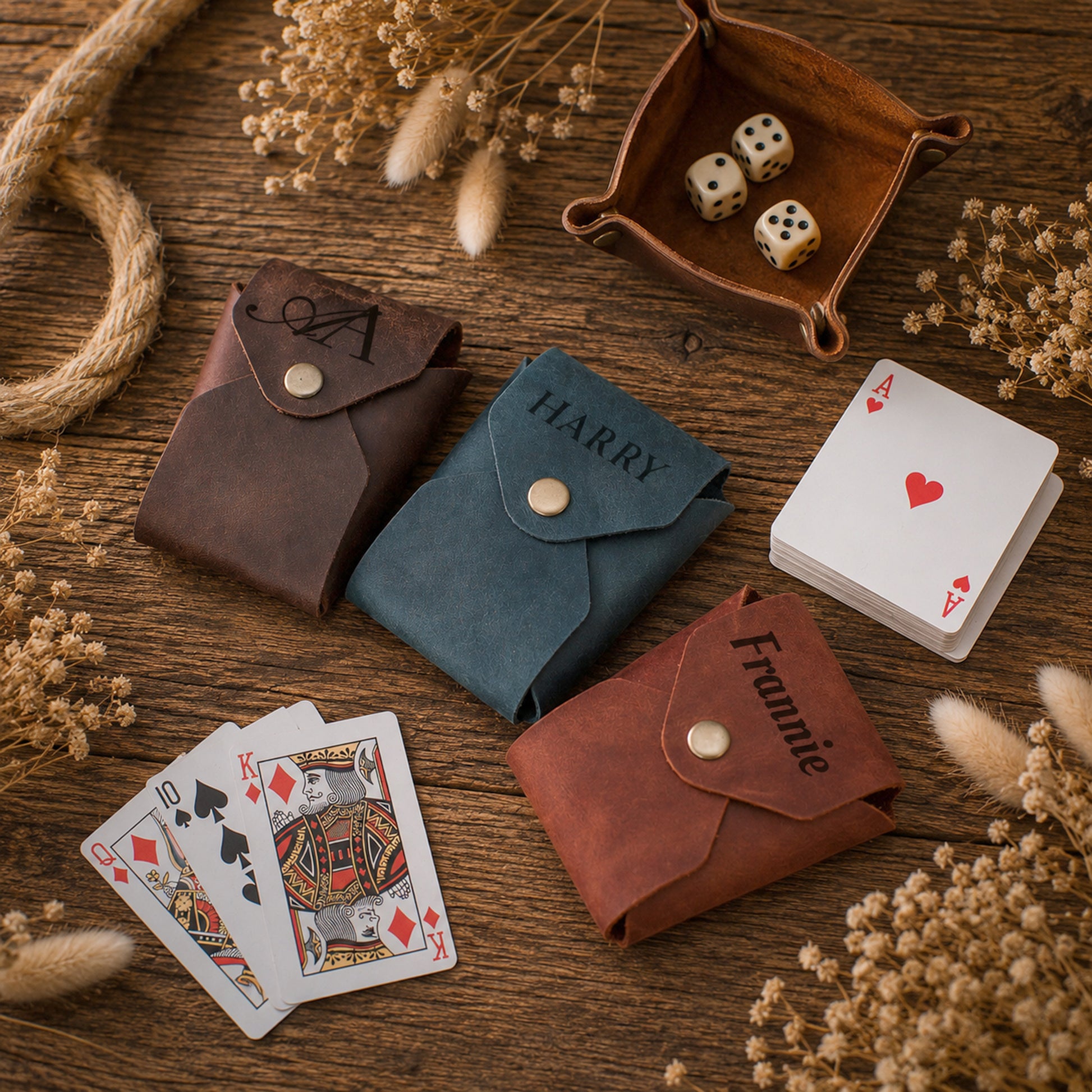 Three personalized leather card sleeves in brown, blue, and reddish-brown, styled on a rustic wooden table with playing cards, dice, and dried flowers.