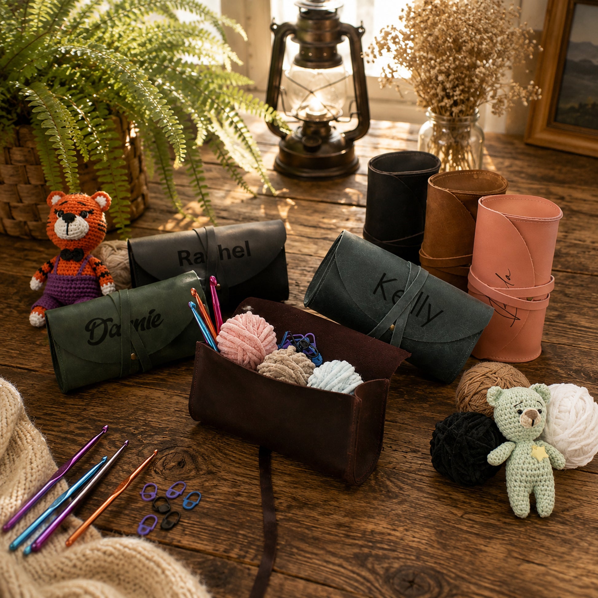A styled flat-lay of personalized crocheting and knitting tools on a rustic wooden table, including three leather pouches personalized with names (Rachel, Kelly, Diane), a dark brown leather organizer, crochet hooks, knitting needles, yarn balls, a small crocheted tiger doll, and a small teddy bear.