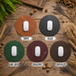 Five round mouse pads in different colors arranged on a rustic wooden surface: Tawny tan, Black, Dark Green, Burgundy, and Chocolate. Each pad is shown with a white computer mouse on top, with the color name printed below.
