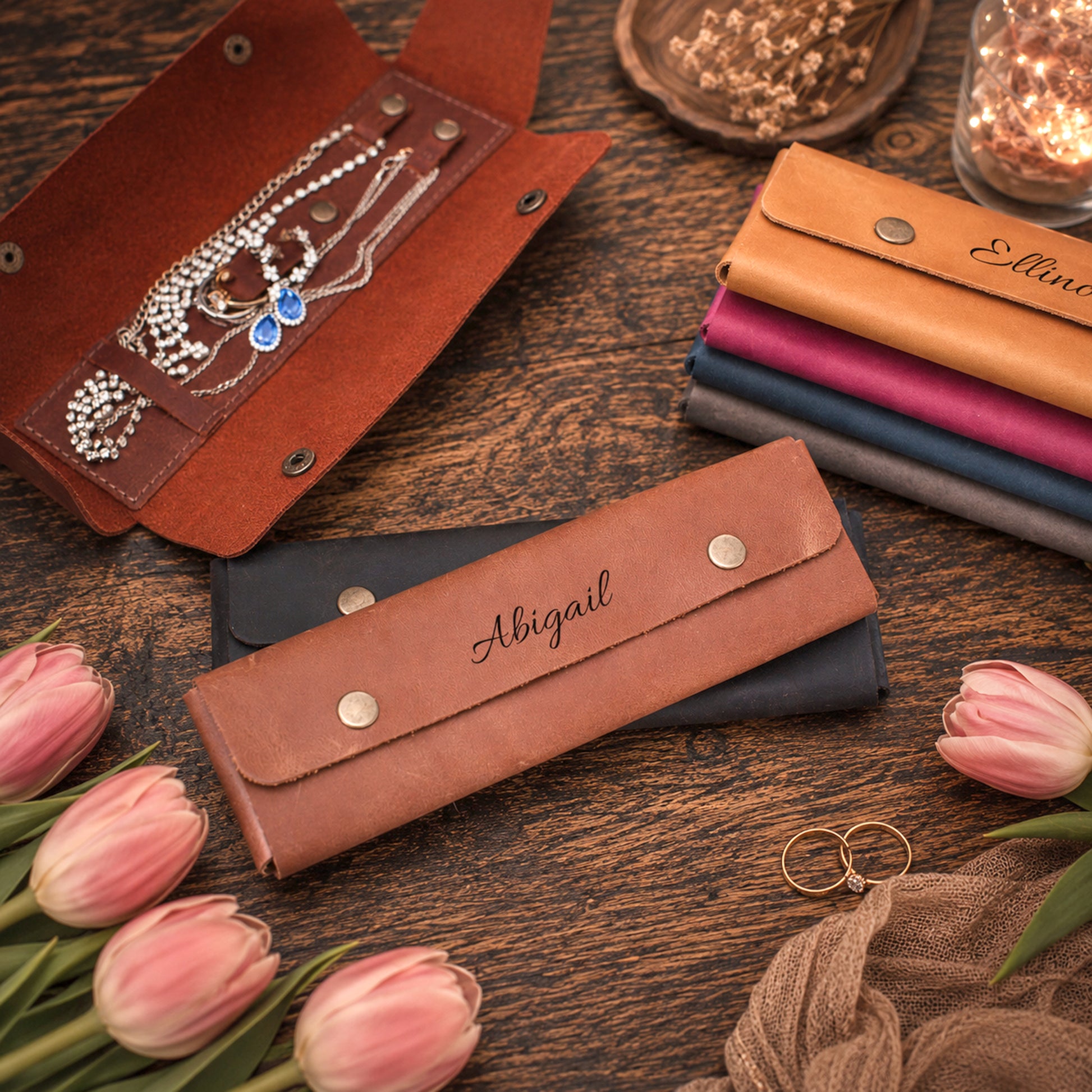 Personalized leather jewelry organizer cases on a rustic wooden table, styled with pink tulips, rings, and other matching cases in different colors.