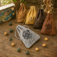 Five personalized drawstring pouches in brown, tan, and gray velvet are arranged on a rustic wooden tabletop with scattered polyhedral gaming dice. The front gray pouch is most prominent, featuring an engraved black mandala and the initials 'P.