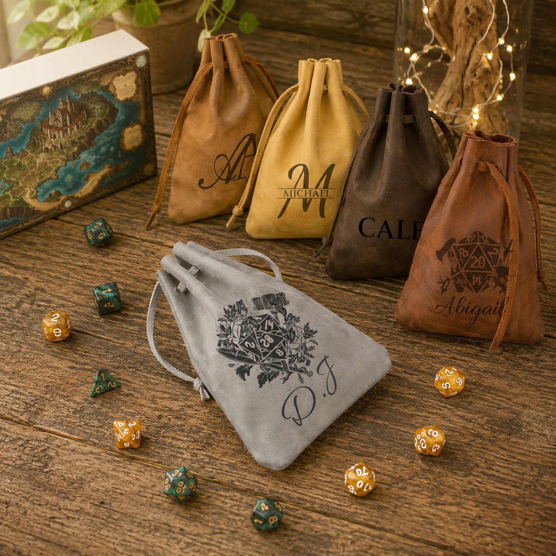 Five personalized drawstring pouches in brown, tan, and gray velvet are arranged on a rustic wooden tabletop with scattered polyhedral gaming dice. The front gray pouch is most prominent, featuring an engraved black mandala and the initials 'P.