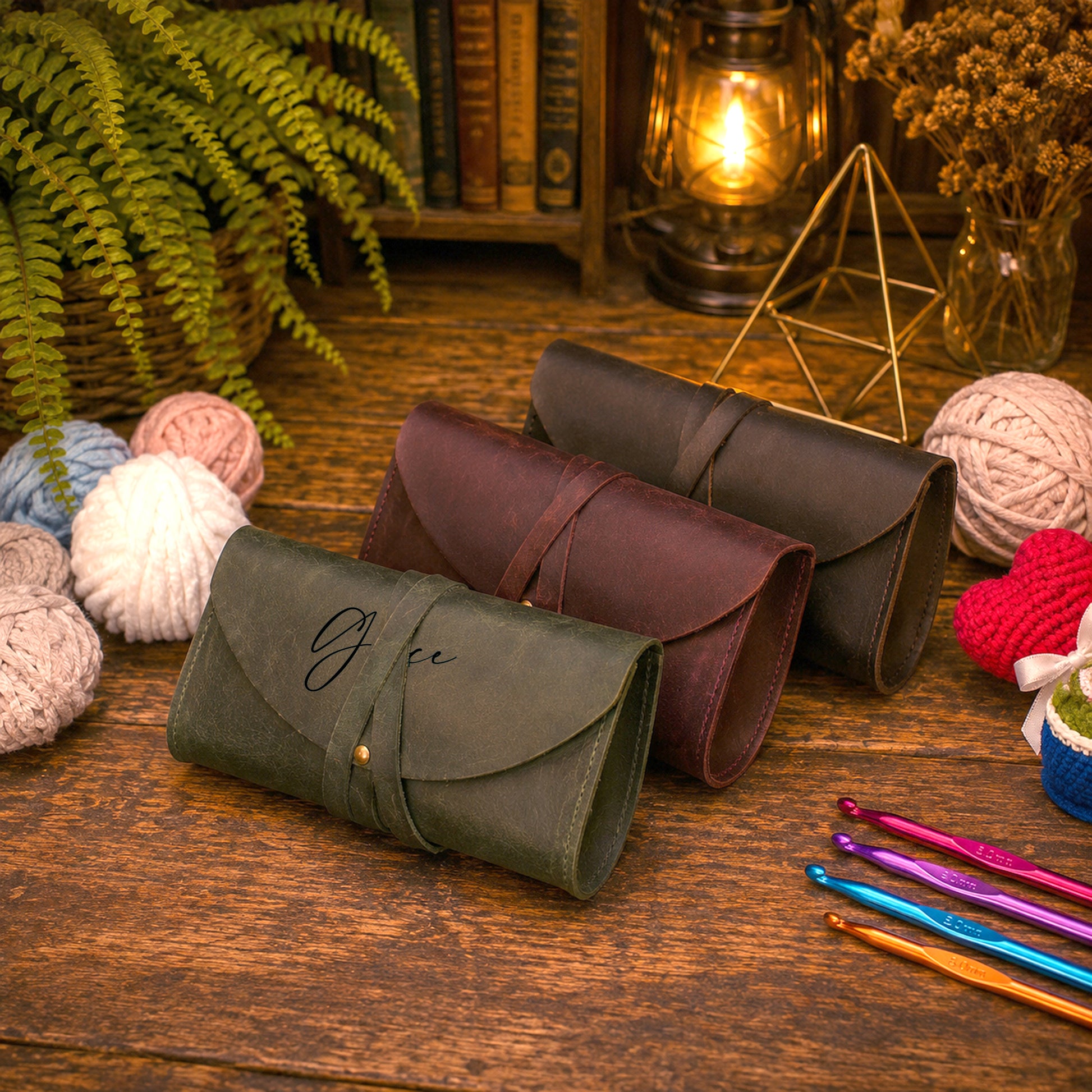 Three leather pouches or cases in olive green, burgundy, and dark brown, arranged on a rustic wooden table with yarn balls, crochet hooks, a plant, and a lantern in the background.