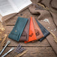 Four small leather pouches or card cases in teal, brown, orange, and dark gray are arranged on a rustic wooden surface with an open book, fabric, dried flowers, beads, and makeup brushes around them.