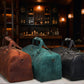 Three leather duffel bags in brown, green, and black on a wooden surface with a bar background.