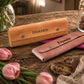 Two personalized leather accessory cases on a rustic wooden table, one tan with the name 'DAMARIS' and one dusty pink with the name 'Frannie', surrounded by pink tulips, a pocket watch, and other decorative props.