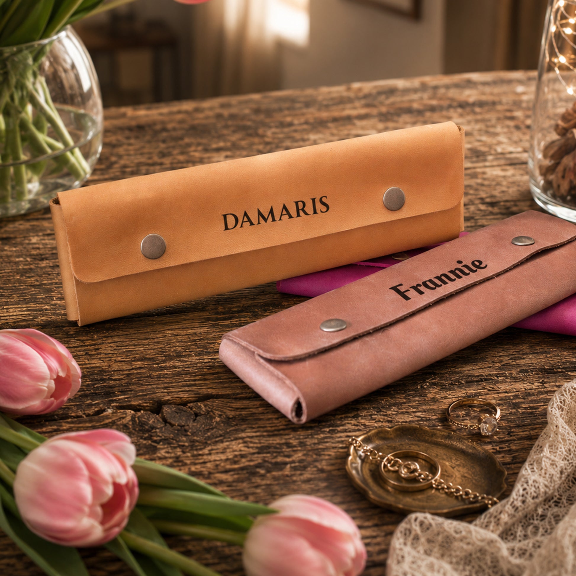 Two personalized leather accessory cases on a rustic wooden table, one tan with the name 'DAMARIS' and one dusty pink with the name 'Frannie', surrounded by pink tulips, a pocket watch, and other decorative props.