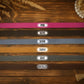 Six faux leather belt straps in different colors are laid out horizontally on a rustic wooden background, each labeled with its color name: PINK, OLIVE, DENIM, TAWNY, GRAY, and CHOCOLATE.