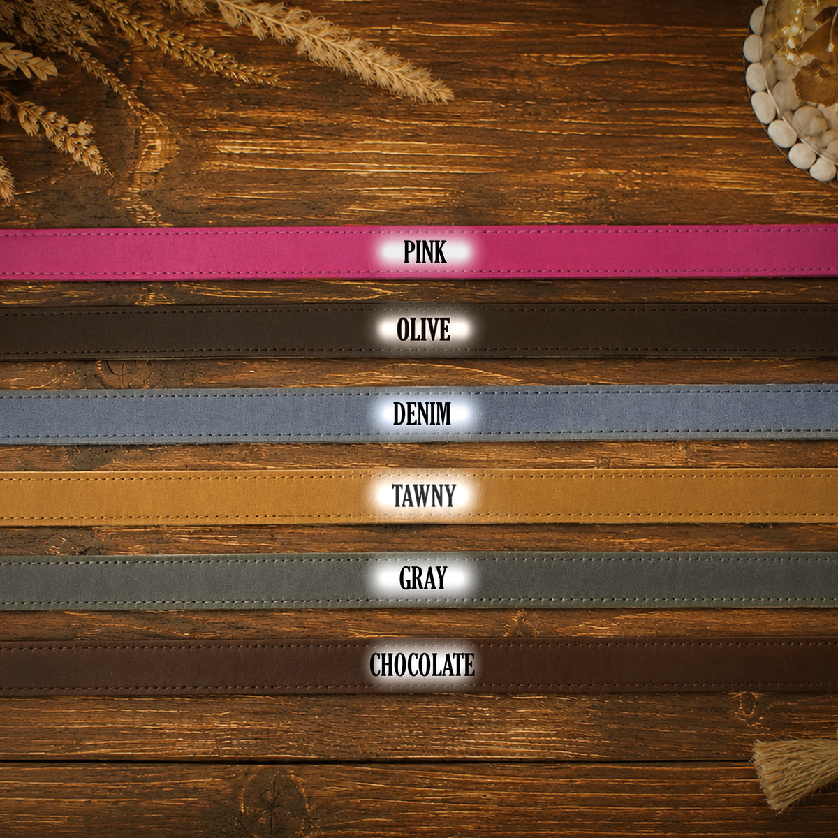 Six faux leather belt straps in different colors are laid out horizontally on a rustic wooden background, each labeled with its color name: PINK, OLIVE, DENIM, TAWNY, GRAY, and CHOCOLATE.
