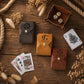 Four leather playing card wallets in dark gray, tan, and brown, arranged on a rustic wooden table with playing cards, dice, and dried flowers.