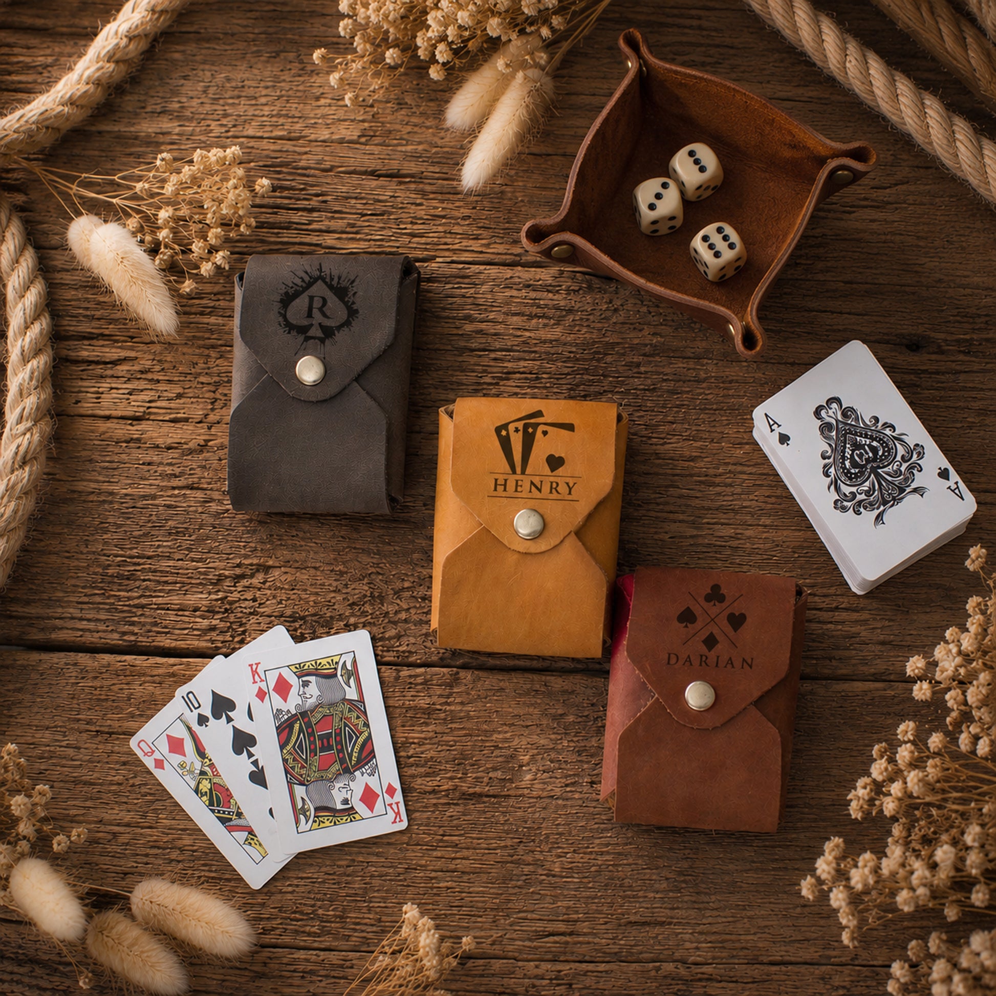 Four leather playing card wallets in dark gray, tan, and brown, arranged on a rustic wooden table with playing cards, dice, and dried flowers.