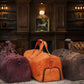 Three duffel bags in purple, orange, and brown on a patterned rug with a wooden panel background.