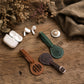 Three personalized leather keychains in brown, green, and dark brown are arranged on a rustic wooden table alongside white wireless earbuds in their charging case, dried flowers, and a piece of linen fabric.