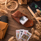 A brown leather playing card holder pouch on a rustic wooden table, shown with playing cards and surrounded by dried flowers, rope, glasses, and a book.