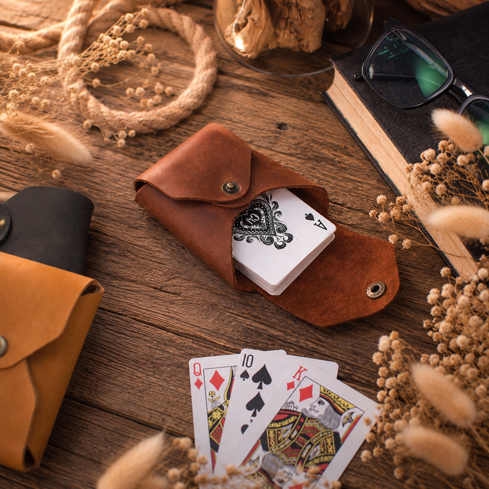 A brown leather playing card holder pouch on a rustic wooden table, shown with playing cards and surrounded by dried flowers, rope, glasses, and a book.