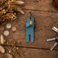 A small blue leather key holder or pouch with a gold snap button and metal key ring, placed on a rustic wooden surface with dried flowers, coins, and keys around it.