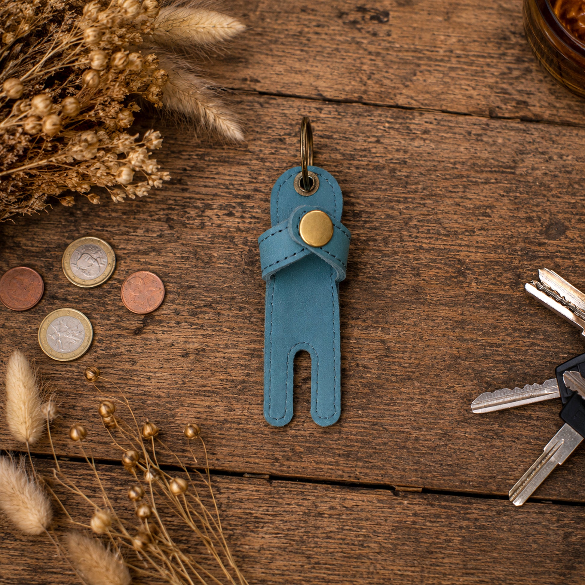 A small blue leather key holder or pouch with a gold snap button and metal key ring, placed on a rustic wooden surface with dried flowers, coins, and keys around it.