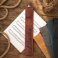 A personalized brown leather music stand sleeve with the name 'Mattin' and monogram 'M' engraved, displayed on a wooden tabletop with sheet music, rope, and other color options.