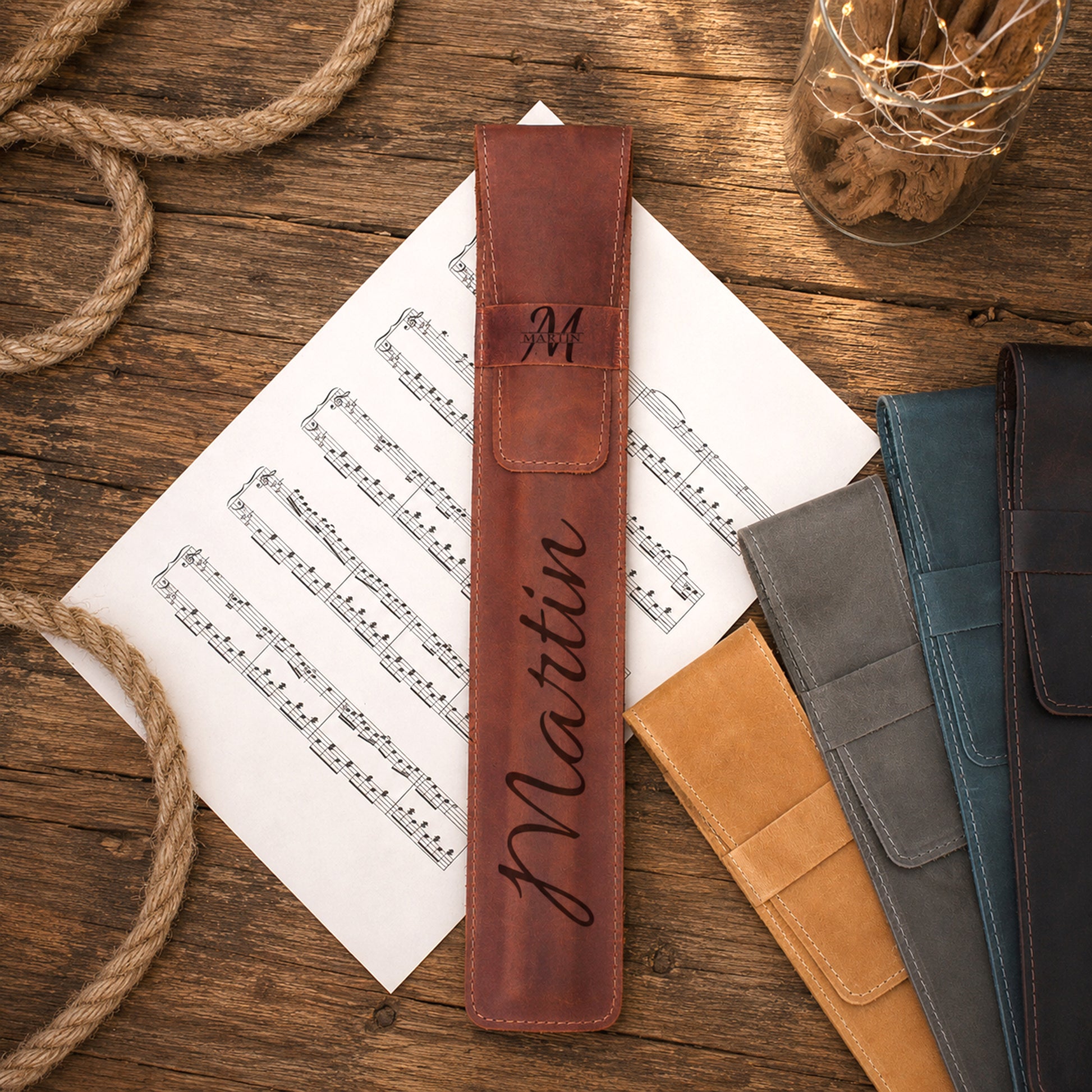 A personalized brown leather music stand sleeve with the name 'Mattin' and monogram 'M' engraved, displayed on a wooden tabletop with sheet music, rope, and other color options.