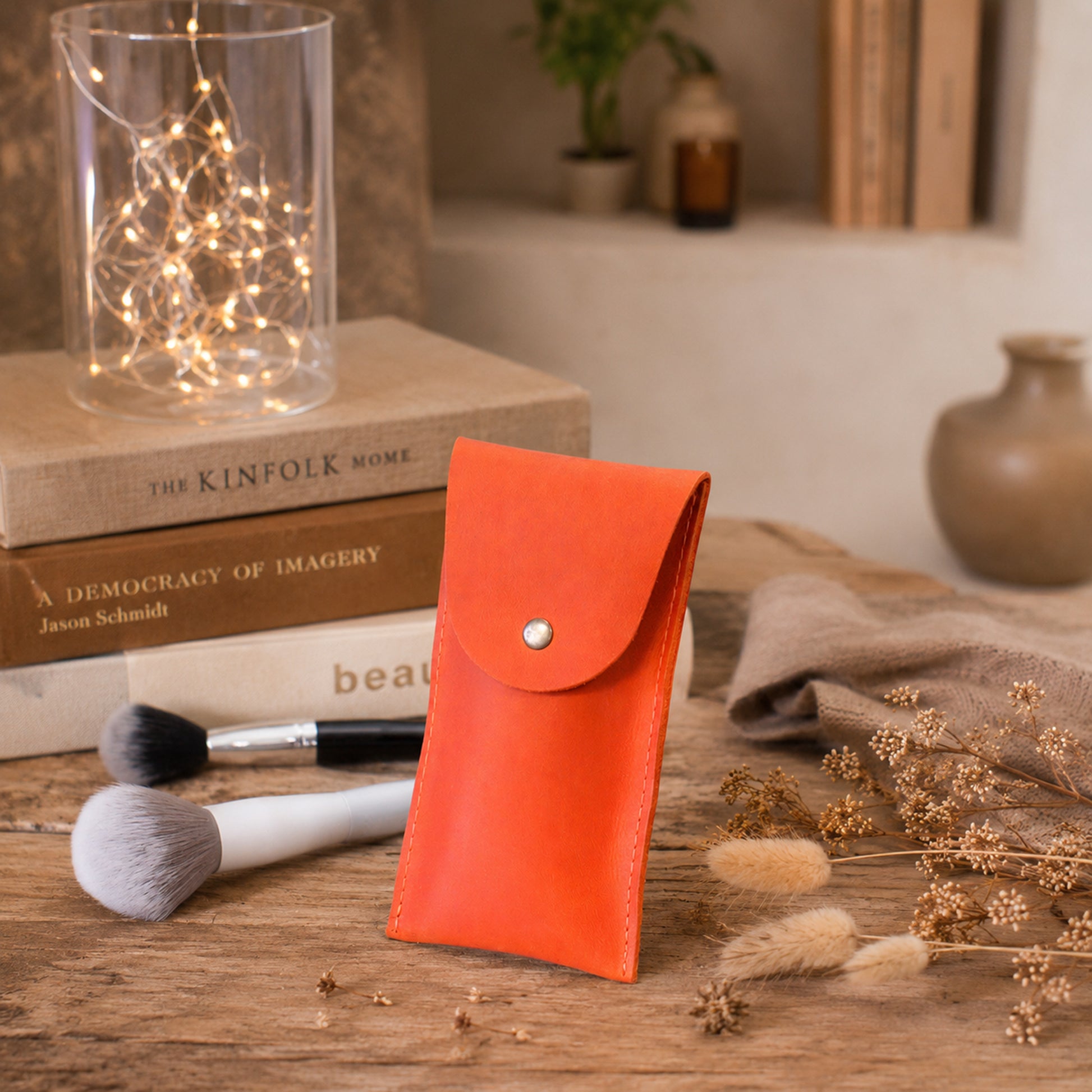 A bright orange leather pouch with a silver snap button, standing on a rustic wooden table surrounded by makeup brushes, dried flowers, and decorative books.