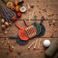 Four leather golf tees in orange, brown, green, and black are arranged on a rustic wooden tabletop with golf tees, dried flowers, a compass, and a golf ball nearby.
