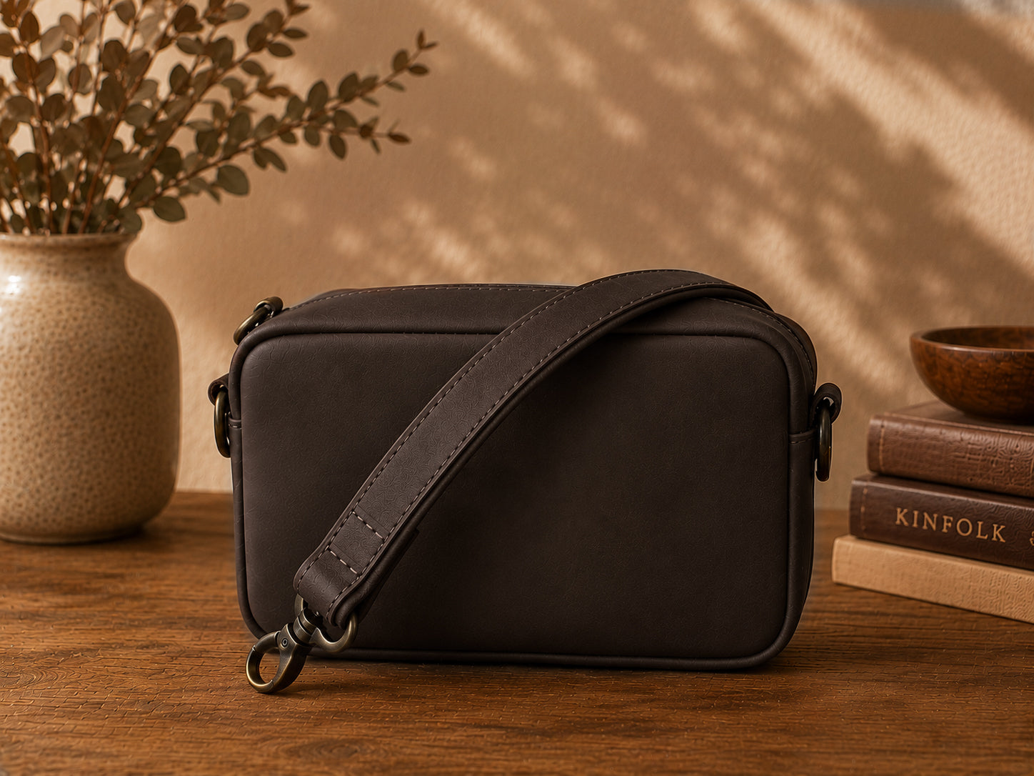Chocolate leather crossbody bag on a wooden surface with a vase and books in the background