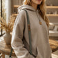 A woman wearing a light beige oversized hoodie with a green shoulder strap and a wooden pendant necklace in a warm, modern living room setting.