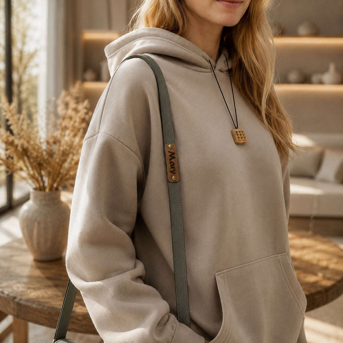 A woman wearing a light beige oversized hoodie with a green shoulder strap and a wooden pendant necklace in a warm, modern living room setting.