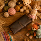 A dark brown leather journal or notebook cover with a wrap-around strap, styled on a rustic wooden surface with crochet hooks, yarn balls, lace, and handmade stuffed toys.