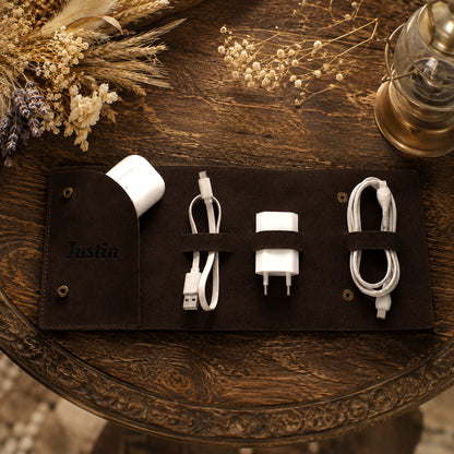 A dark brown personalized roll-up leather charging cable organizer on a rustic wooden table, holding a white wireless earbud case, a USB-C cable, a white power adapter, and another coiled cable. The organizer is personalized with the name 'Justin' in black script.