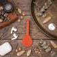 A terracotta orange leather luggage tag with a matching leather strap and silver metal buckle, styled in a flat-lay arrangement on a rustic wooden table with various decorative props including a leather case, AirPods, a compass, and dried botanicals.