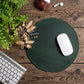 A dark green round leather mouse pad on a rustic wooden desk, styled with a white wireless mouse, a keyboard corner, dried flowers, and a plant.