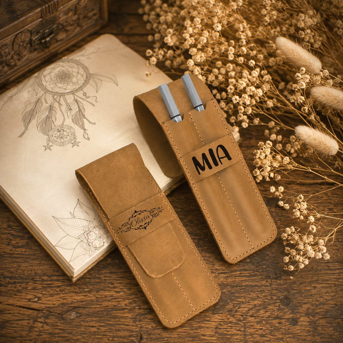 Two tan leather pen holders on a rustic wooden table, styled with dried flowers, a sketchbook, and a wooden box. One holder is open, holding two silver pens, and is personalized with the name 'MIA' stitched on the front.