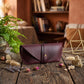 A burgundy leather envelope-style clutch or case with a single strap and a small gold button, placed on a rustic wooden table in a warm, styled interior setting with dried flowers, a plant, and decorative objects.
