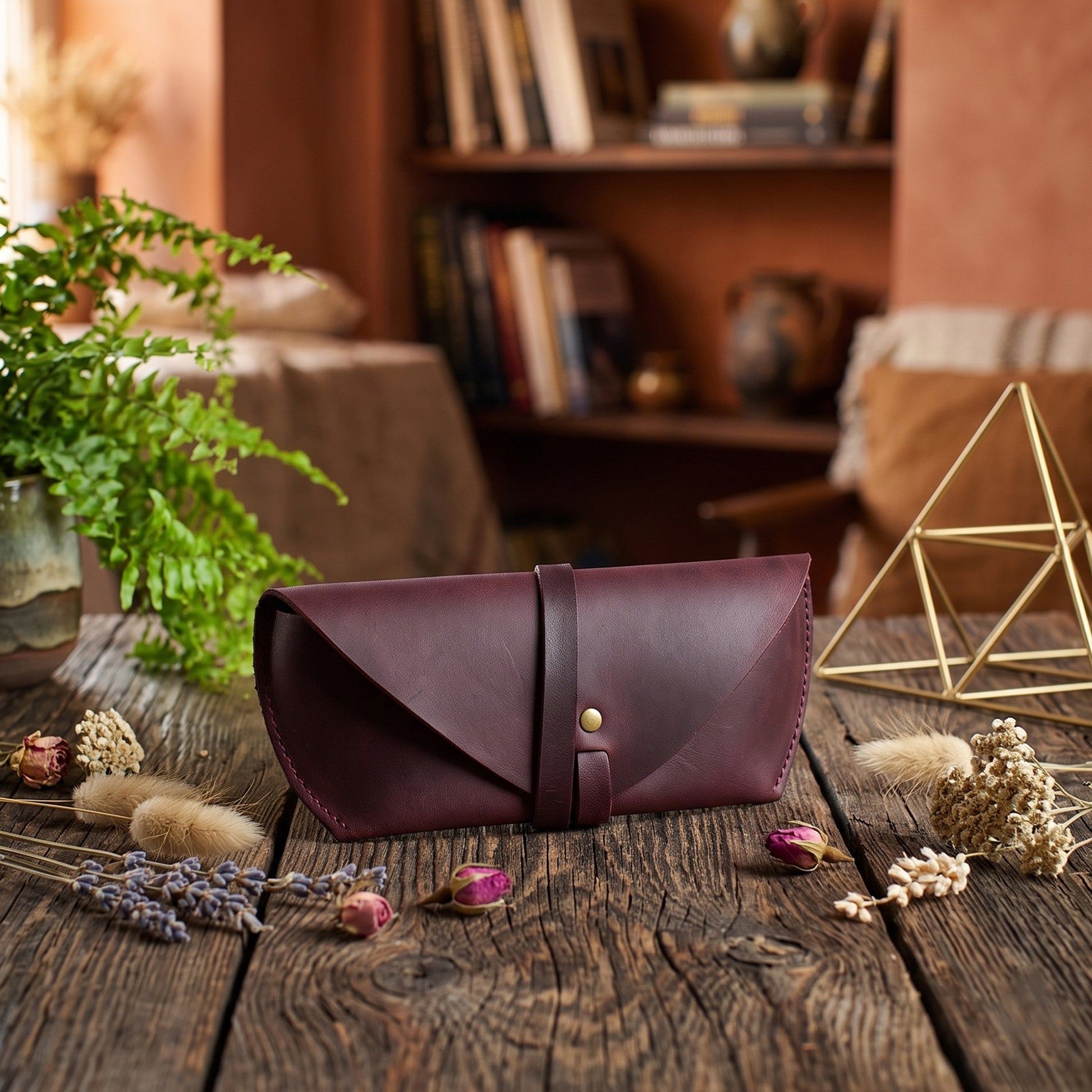 A burgundy leather envelope-style clutch or case with a single strap and a small gold button, placed on a rustic wooden table in a warm, styled interior setting with dried flowers, a plant, and decorative objects.
