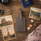 A gray leather luggage tag lies on a rustic wooden tabletop, surrounded by travel-themed props including postcards, a passport, coins, a camera lens, a compass, and a map. No person or animal is present, but the styled scene is not a plain studio product shot; however, there is no real environment being depicted as the main context beyond the tabletop styling, so this fits Product/Packaging rather than Lifestyle.