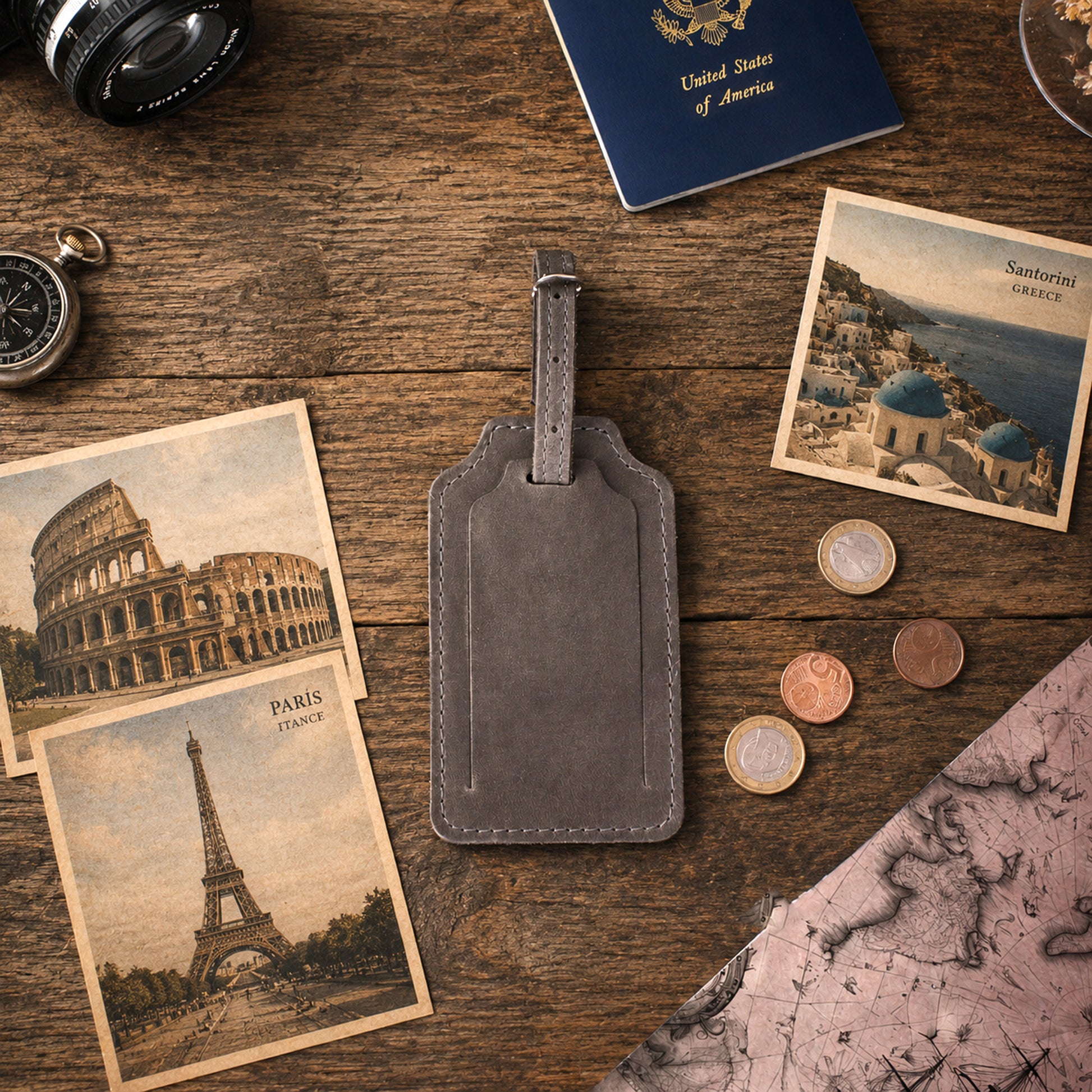 A gray leather luggage tag lies on a rustic wooden tabletop, surrounded by travel-themed props including postcards, a passport, coins, a camera lens, a compass, and a map. No person or animal is present, but the styled scene is not a plain studio product shot; however, there is no real environment being depicted as the main context beyond the tabletop styling, so this fits Product/Packaging rather than Lifestyle.