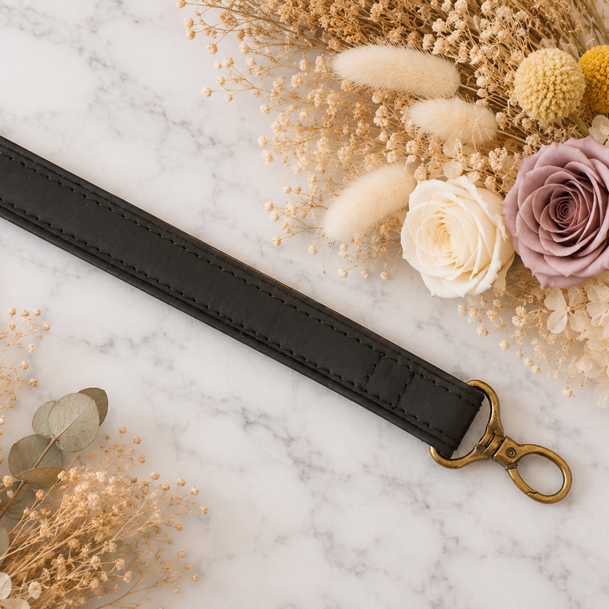 Black leather strap with gold clasp on a marble surface with flowers and dried plants.