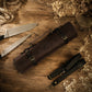 A dark brown leather chef's knife roll with black straps and buckles, laid on a rustic wooden table with two chef knives, dried flowers, and rope props.