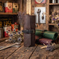 A dark brown leather roll-up tool pouch with a black strap and brass buckle stands on a rustic wooden table, surrounded by vintage-style tools, dried flowers, and other leather tool rolls in a styled workshop setting.