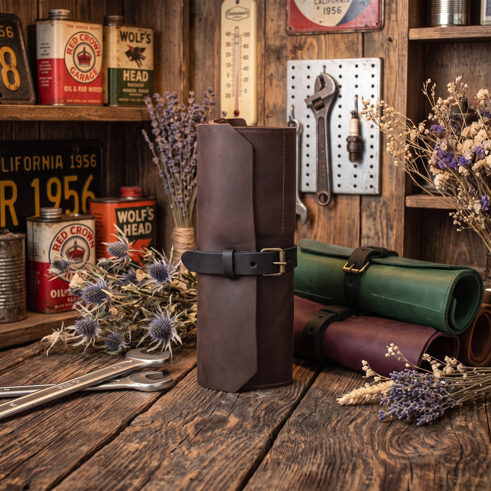 A dark brown leather roll-up tool pouch with a black strap and brass buckle stands on a rustic wooden table, surrounded by vintage-style tools, dried flowers, and other leather tool rolls in a styled workshop setting.