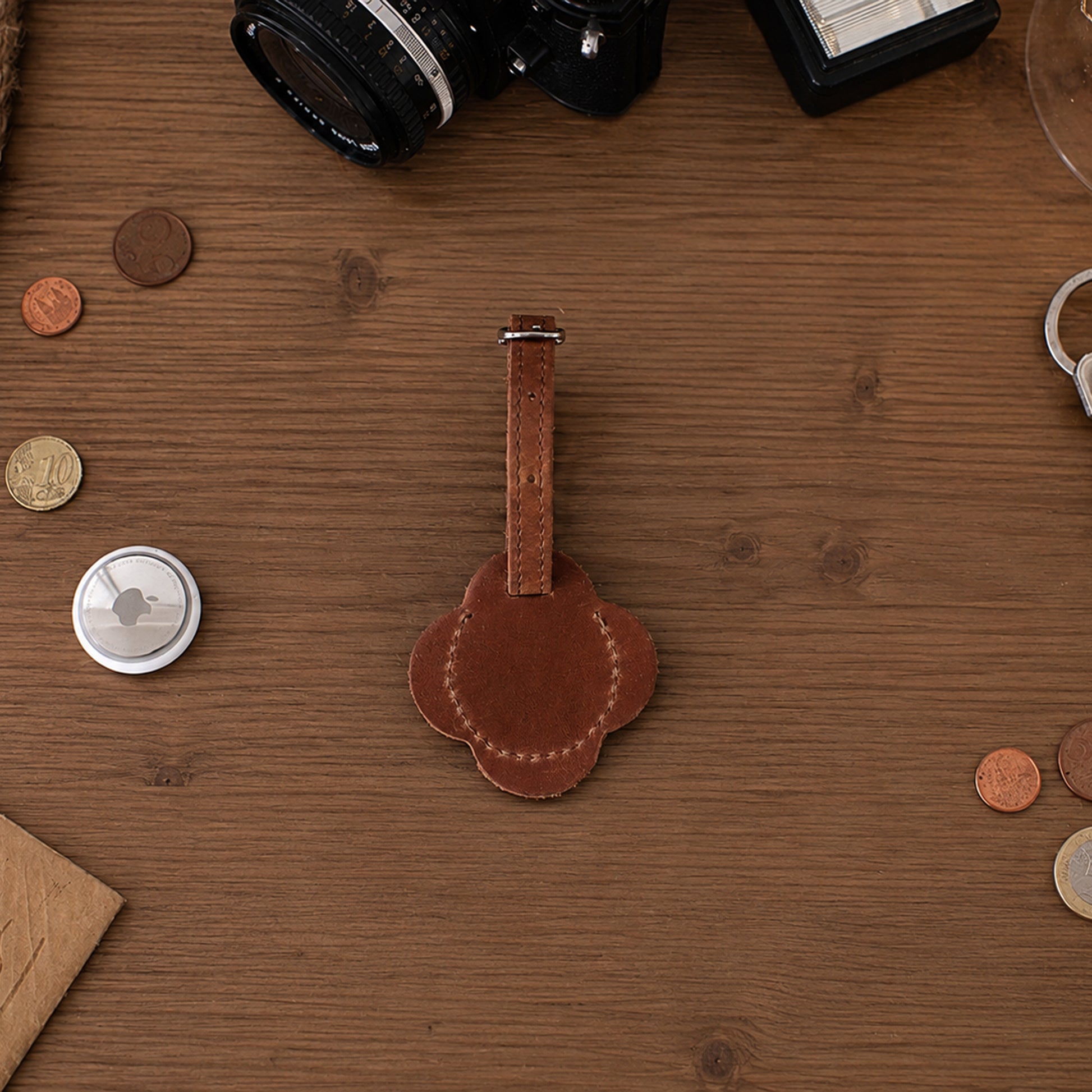 A brown leather luggage tag with a matching strap, laid flat on a wooden tabletop surrounded by small props like coins, a camera lens, and a small round accessory.