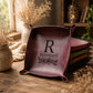 A square leather tray with rolled corners and a dark monogram 'R Rosemary' printed on the front, displayed on a rustic wooden table with dried flowers and a stack of similar trays in the background.