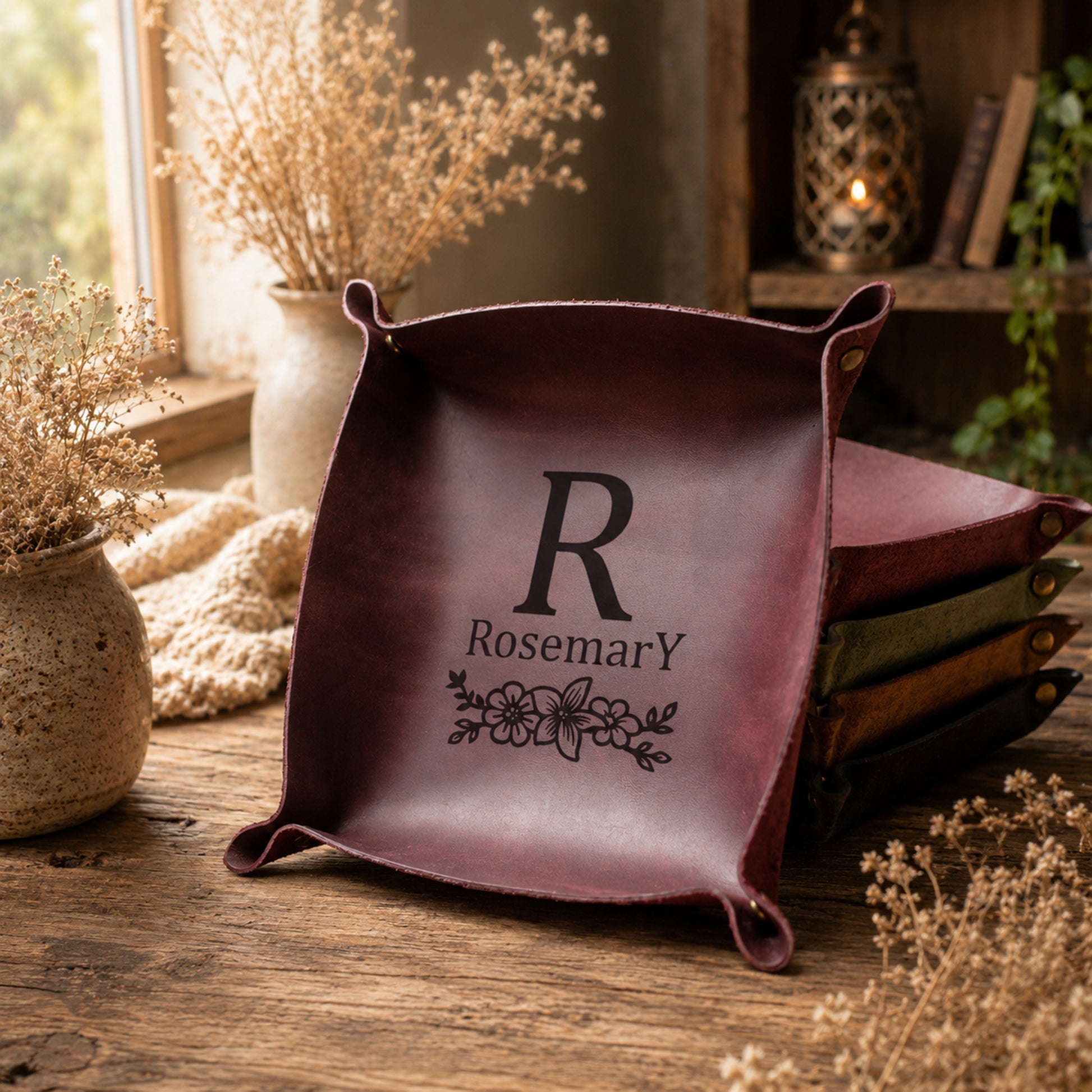 A square leather tray with rolled corners and a dark monogram 'R Rosemary' printed on the front, displayed on a rustic wooden table with dried flowers and a stack of similar trays in the background.
