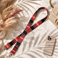 Red and black striped strap with gold hardware on a light surface with a phone and dried flowers.