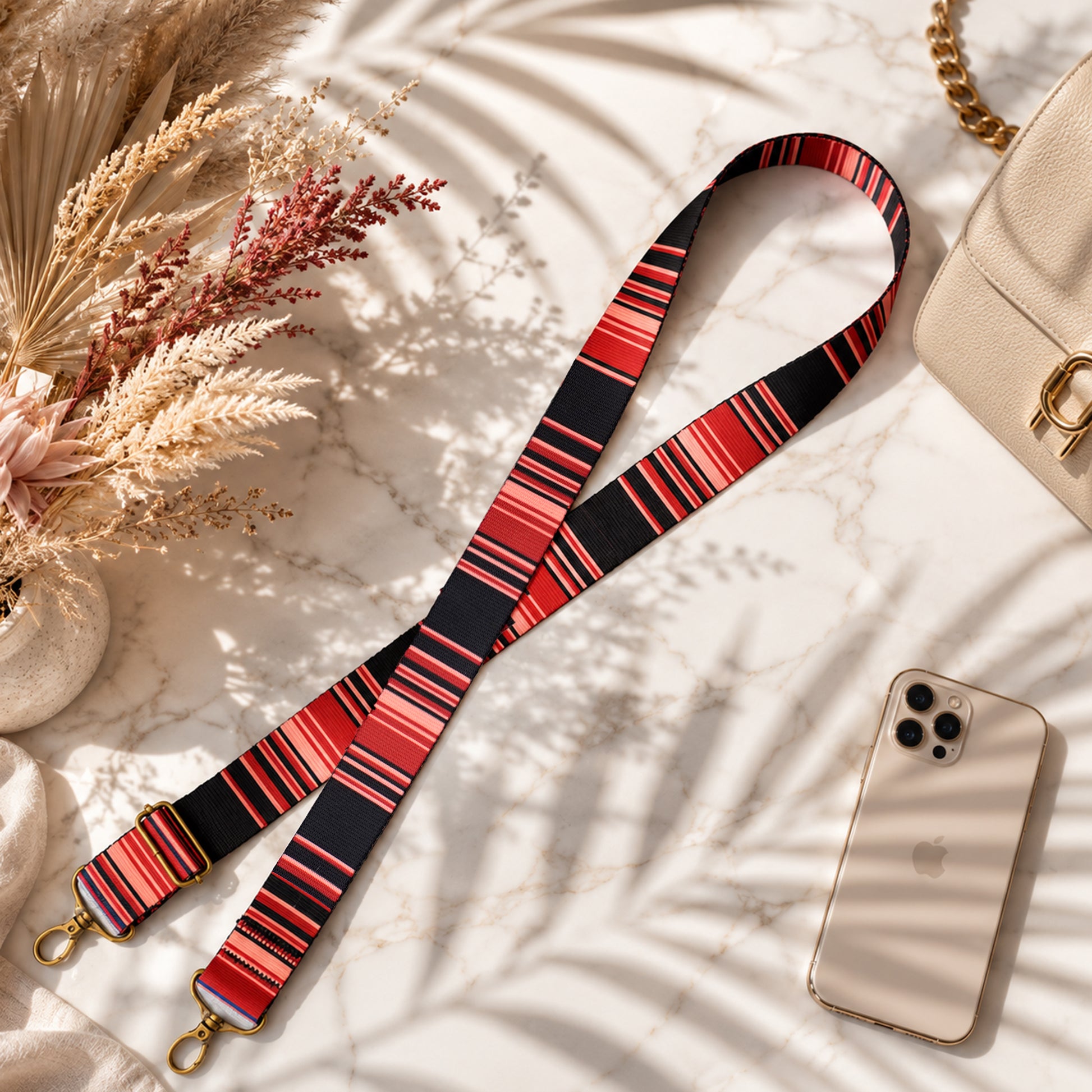 Red and black striped strap with gold hardware on a light surface with a phone and dried flowers.