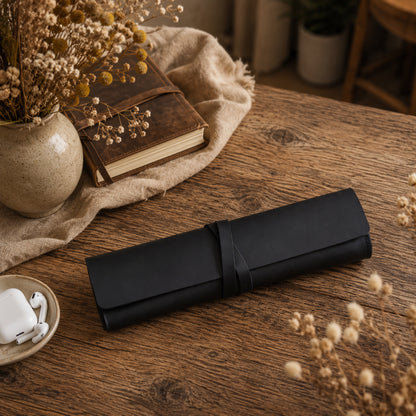 A black rolled leather or faux-leather case with a wrap strap sits on a rustic wooden table, surrounded by dried flowers, a small bowl of white earbuds, and a vintage-style book.