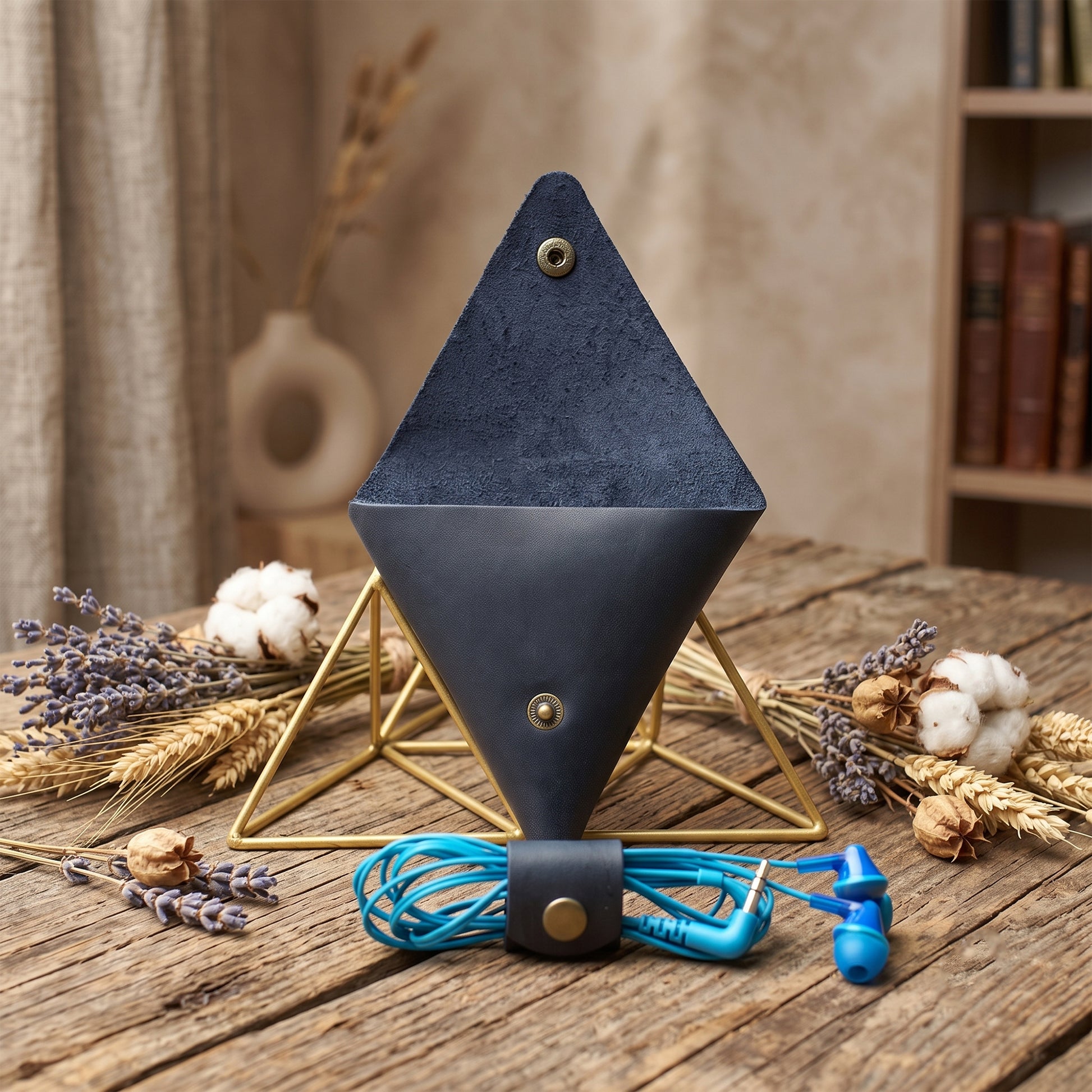 A dark blue cone-shaped speaker or speaker component displayed on a gold metal geometric stand, with blue wired earphones resting in front on a rustic wooden table. The scene is styled with dried lavender, wheat, and cotton in a warm, indoor setting.