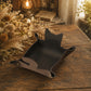 A dark brown leather cat-shaped tray with a small metal snap button, resting on a rustic wooden table with dried flowers and a warm lantern in the background.